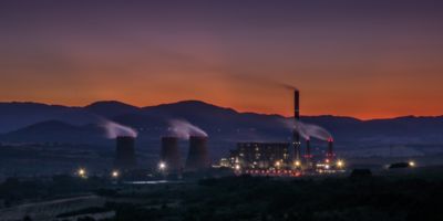 Picture of an industrial site at sunset, emitting clouds, with mountains in the background.