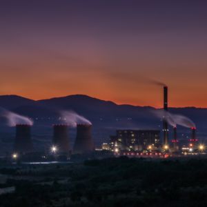 Picture of an industrial site at sunset, emitting clouds, with mountains in the background.